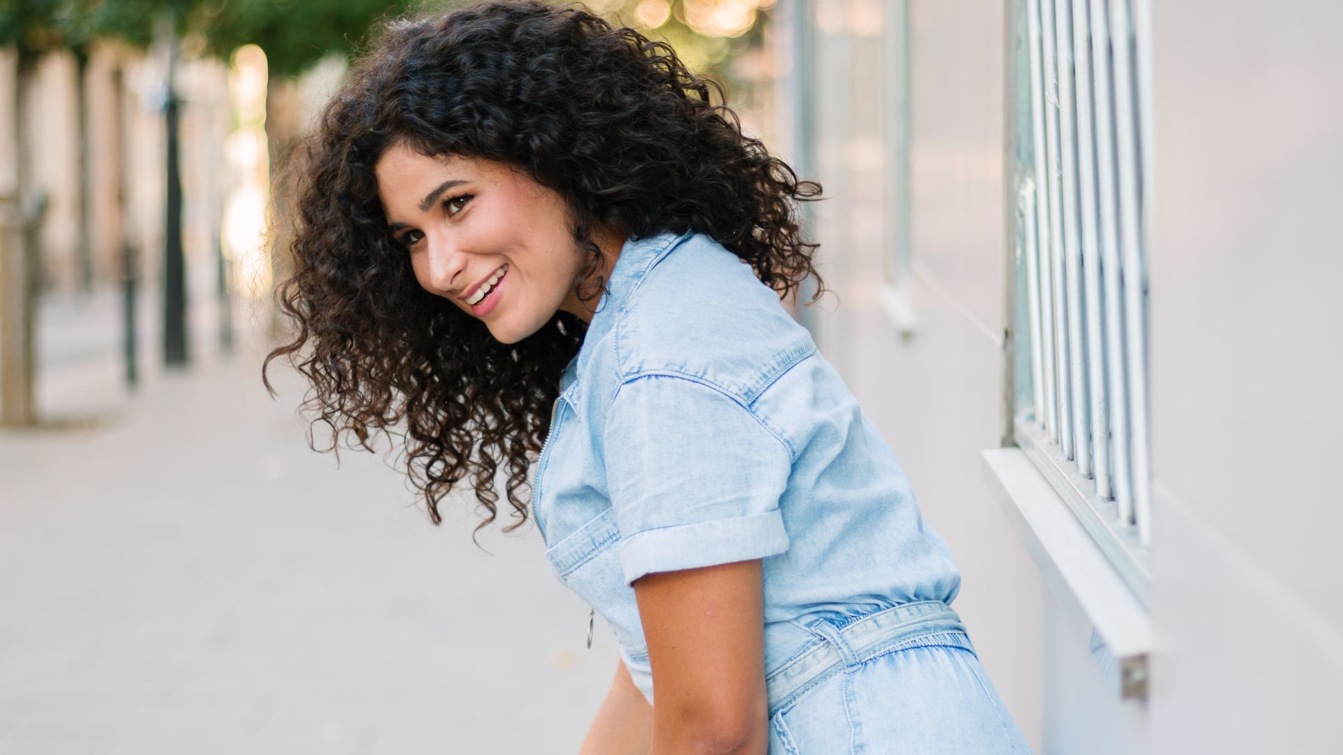Mulher sorridente com cabelo encaracolado a posar ao ar livre com um fato de ganga azul.